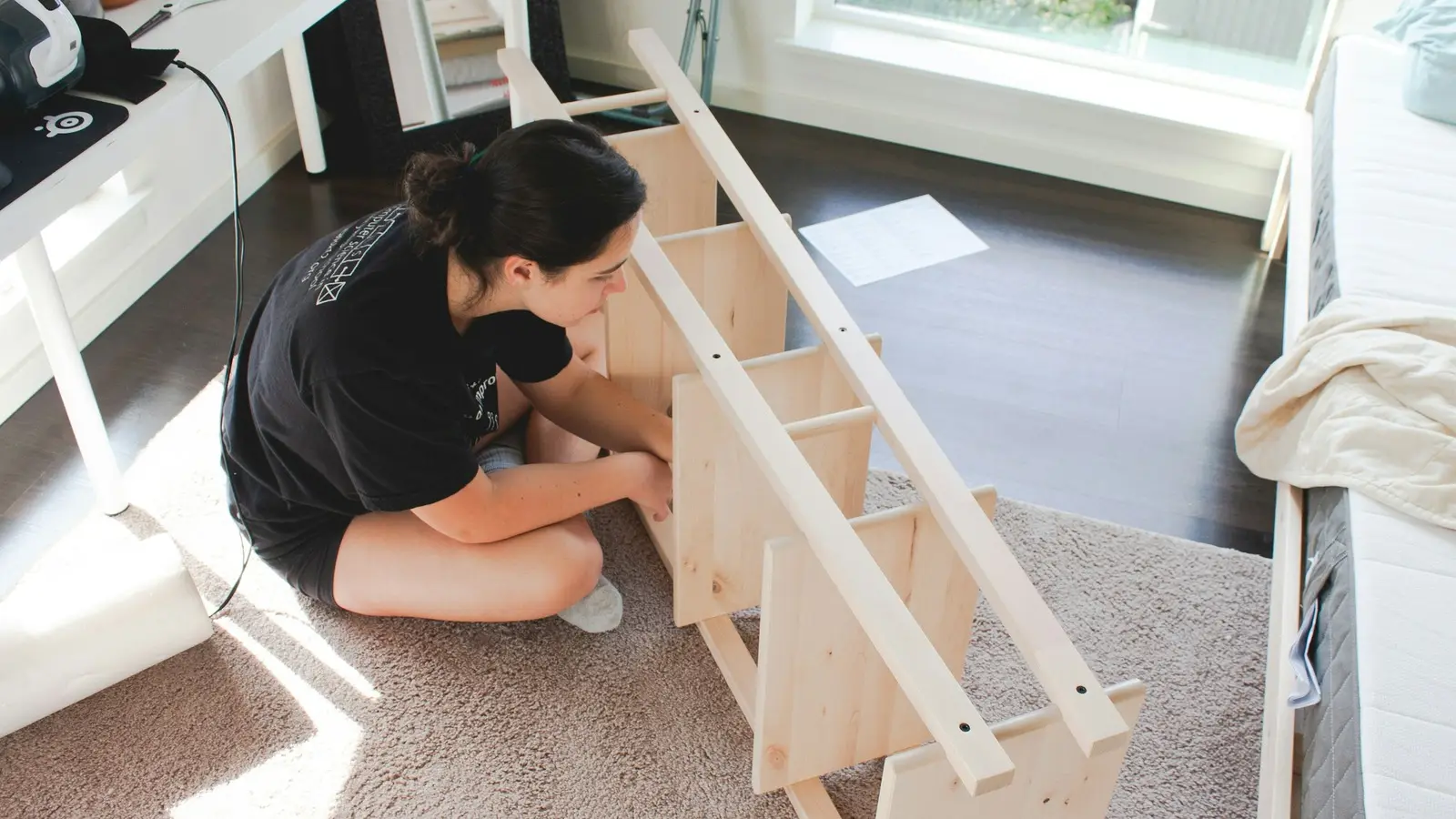 Person assembling a flat-pack wooden shelf on the floor of a bright room
